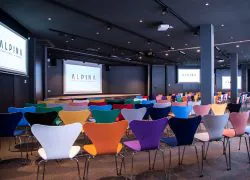 Salle de conférence avec rangées de chaises en plastique moulé multicolores aux pieds chromés face à une scène surélevée, trois écrans muraux affichant le logo de l'hôtel « Alpina » à l'avant, spots encastrés et vidéoprojecteur au plafond sombre au-dessus, une grande salle configurée pour une location de salle.