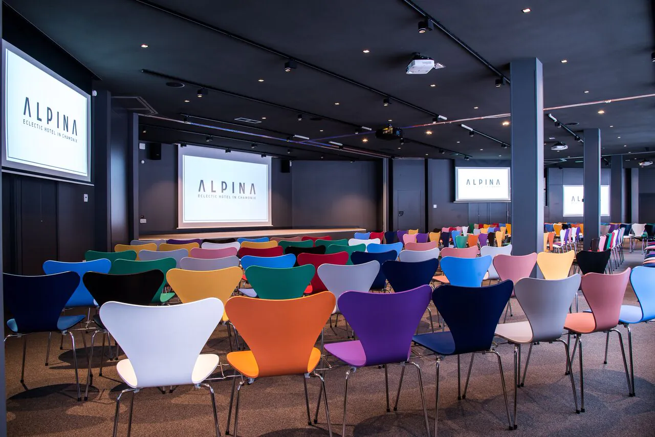 Salle de conférence avec rangées de chaises en plastique moulé multicolores aux pieds chromés face à une scène surélevée, trois écrans muraux affichant le logo de l'hôtel « Alpina » à l'avant, spots encastrés et vidéoprojecteur au plafond sombre au-dessus, une grande salle configurée pour une location de salle.