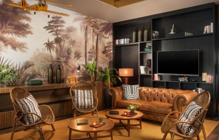 Salon d'hôtel avec canapé chesterfield en cuir marron et fauteuils en rotin aux coussins à motifs autour de tables basses rondes en bois au premier plan, télévision murale intégrée dans un meuble noir garni de livres et objets décoratifs, fresque murale de jungle avec palmiers et maquette d'avion suspendue à gauche, et buffet en bois avec plantes et bouteilles sous la fresque, un salon adapté à un afterwork.