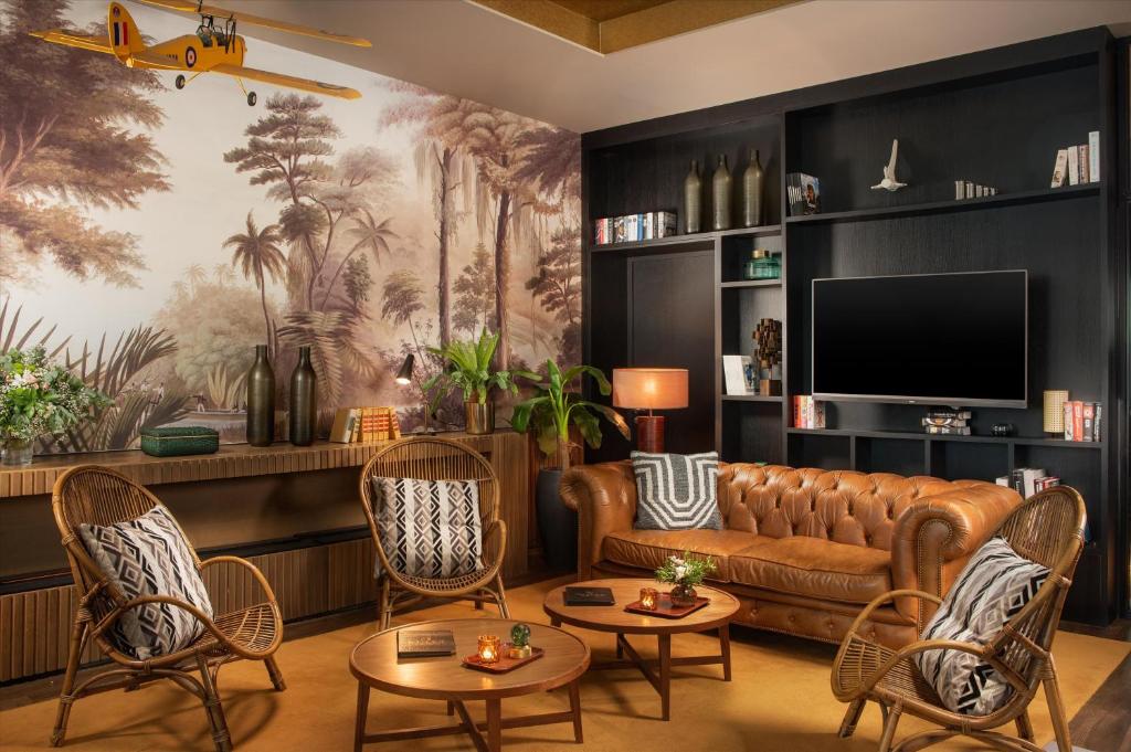 Hotel lounge with a brown leather chesterfield sofa and rattan armchairs with patterned cushions around round wooden coffee tables in the foreground, a wall-mounted television within a black shelving unit displaying books and decorative objects, a palm tree jungle mural with a suspended model aircraft on the left, and a wooden sideboard with plants and bottles beneath the mural, a lounge suited for an afterwork.