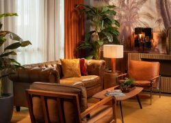 Salon d'hôtel avec canapé chesterfield en cuir marron et fauteuils aux coussins orange et bordeaux autour d'une table basse en bois au premier plan, plantes en pot hautes à gauche, fresque murale de jungle avec sculptures décoratives murales et vase en laiton sur un buffet en bois en arrière-plan, et lampe de table près du canapé, un salon adapté à un afterwork.