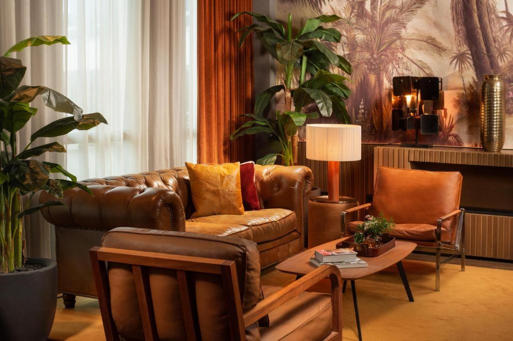 Salon d'hôtel avec canapé chesterfield en cuir marron et fauteuils aux coussins orange et bordeaux autour d'une table basse en bois au premier plan, plantes en pot hautes à gauche, fresque murale de jungle avec sculptures décoratives murales et vase en laiton sur un buffet en bois en arrière-plan, et lampe de table près du canapé, un salon adapté à un afterwork.