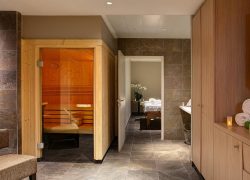 Espace de séminaire bien-être d'hôtel, avec une cabine de sauna en bois à gauche, un couloir menant à une salle de massage au centre, et des rangements en bois avec des serviettes blanches roulées et des bougies à droite, sur un sol carrelé gris.
