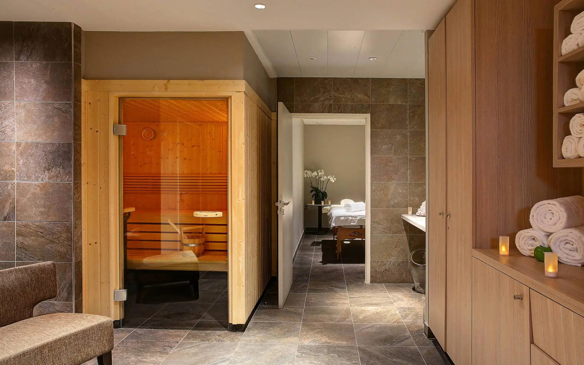 Hotel wellness seminar area, with a wooden sauna cabin on the left, a corridor leading to a massage room in the centre, and wood storage units with rolled white towels and candles on the right, on grey tiled flooring.