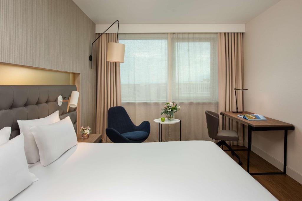 Chambre d'hôtel avec lit aux draps blancs et tête de lit rembourrée grise équipée d'une liseuse murale au premier plan, fauteuil rembourré bleu près d'une table d'appoint ronde avec fleurs, lampadaire derrière eux, et bureau en bois avec chaise pivotante beige près de fenêtres rideautées en arrière-plan, une chambre adaptée à un hôtel pour séminaire.