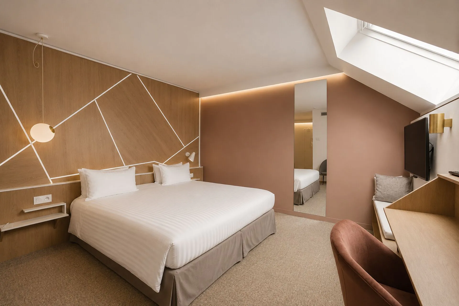 Attic hotel room for group accommodation with a white striped bed against a wood-panelled headboard wall lined with illuminated geometric lines on the left, a terracotta velvet armchair and wooden desk in the foreground on the right, a full-length mirror against a dusty pink wall reflecting the room, and a wall-mounted television beside a skylight above under the sloped ceiling.