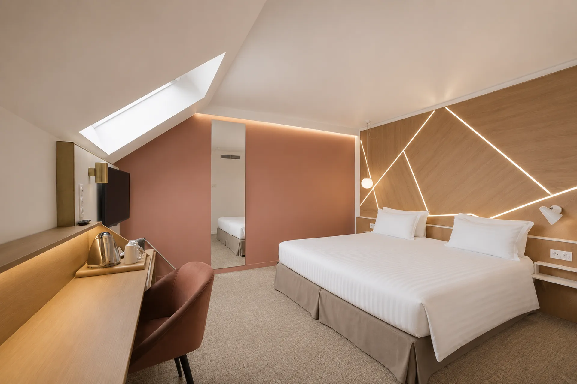 Attic hotel room for group accommodation with a white striped bed under a sloped wood-panelled wall lit by illuminated geometric lines on the right, a terracotta velvet chair and wooden desk with kettle and mug in the foreground on the left, a full-length mirror against a dusty pink wall reflecting the bed, and a skylight above under the sloped ceiling.