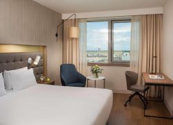 Chambre d'hôtel avec lit aux draps blancs et tête de lit rembourrée grise équipée d'une liseuse murale au premier plan, fauteuil rembourré bleu près d'une table d'appoint ronde avec fleurs, et bureau en bois avec chaise pivotante beige près d'une fenêtre donnant sur une tour de contrôle d'aéroport en arrière-plan, une chambre adaptée à un hôtel pour séminaire.