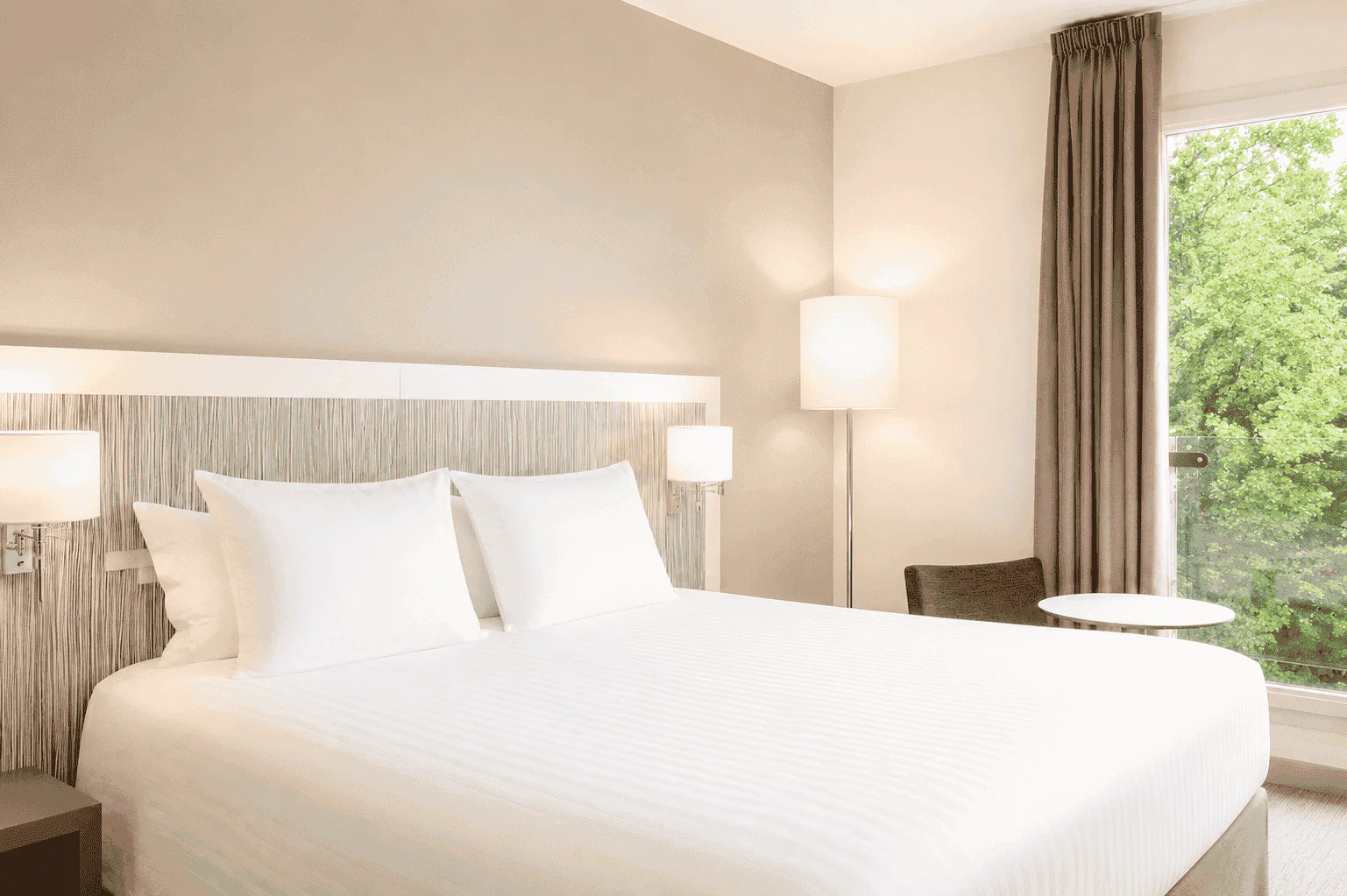 Hotel room for group accommodation, with a large white double bed in the foreground, a wood-effect headboard with wall-mounted reading lights on the left, a floor lamp, armchair and round side table beside a window with grey curtains on the right, and trees visible outside.