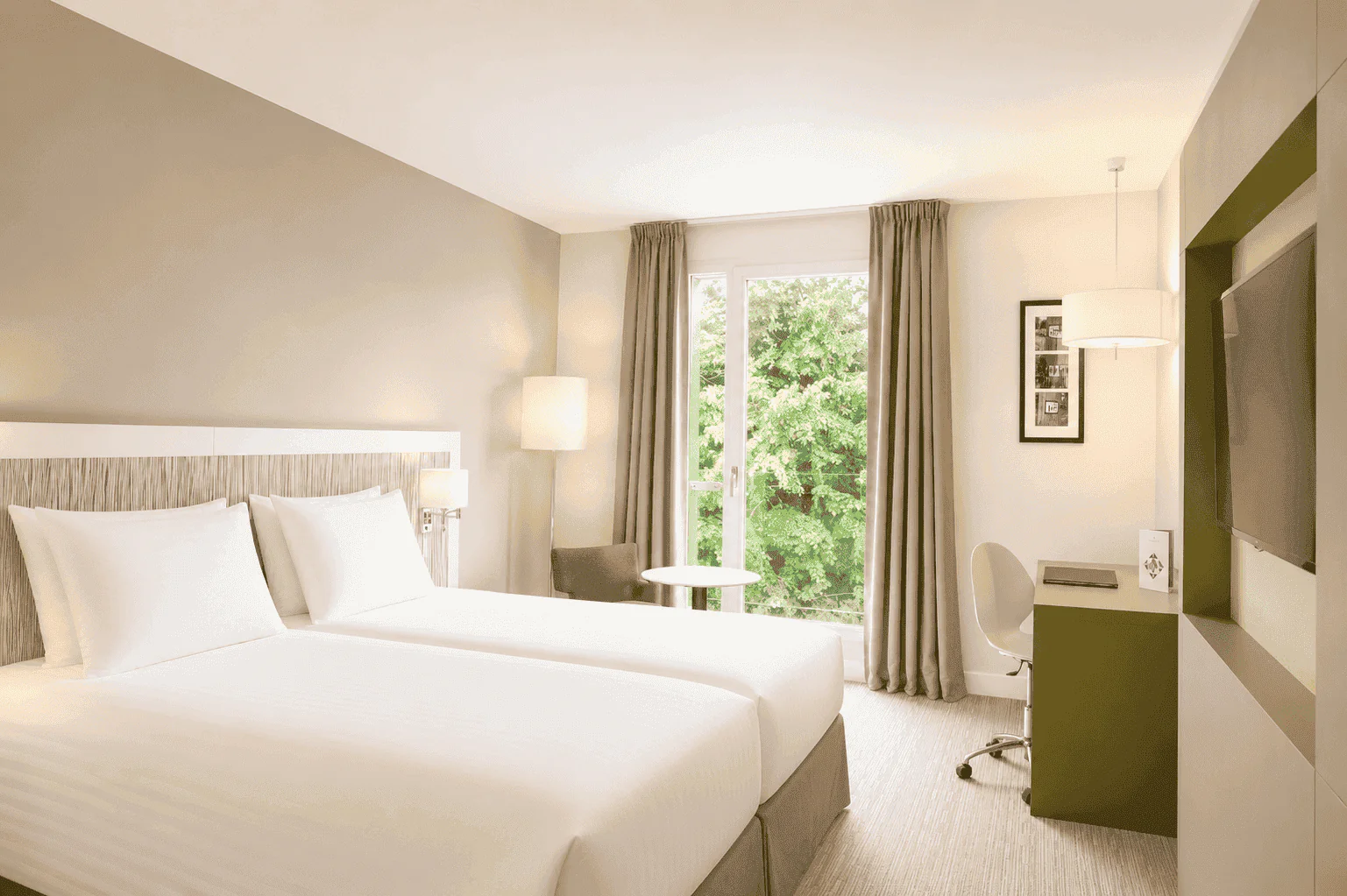 Hotel room for group accommodation, with two white single beds in the foreground on the left, a round table and armchair beside a window with grey curtains in the centre, a desk with chair and wall-mounted television on the right, and trees visible outside.