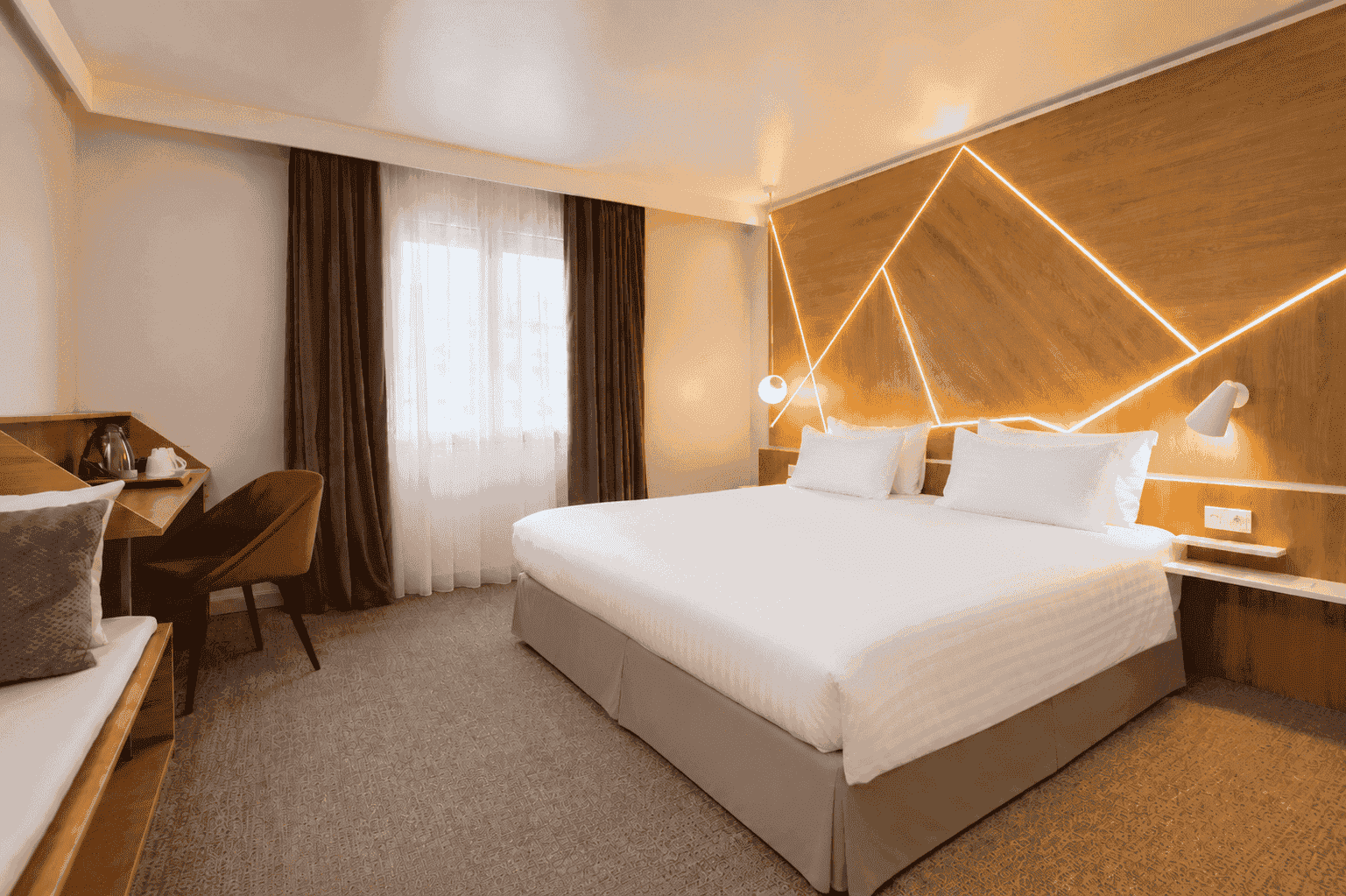 Hotel room for group accommodation, with a large double bed on the right in the foreground, a wood headboard with illuminated geometric lines on the wall, a desk with chair and hospitality tray on the left, and a window with sheer and dark curtains in the background on grey carpet.