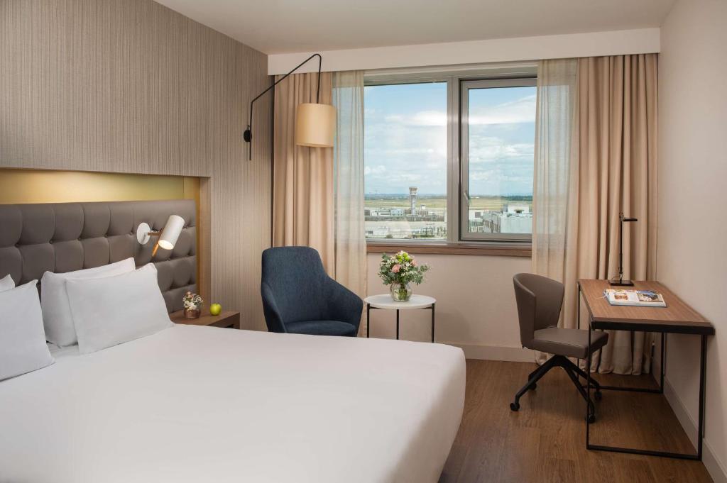 Chambre d'hôtel avec lit aux draps blancs et tête de lit rembourrée grise équipée d'une liseuse murale au premier plan, fauteuil rembourré bleu près d'une table d'appoint ronde avec fleurs, et bureau en bois avec chaise pivotante beige près d'une fenêtre donnant sur une tour de contrôle d'aéroport en arrière-plan, une chambre adaptée à un hôtel pour séminaire.