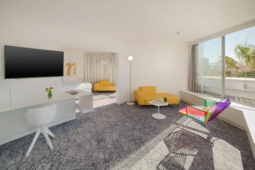 Suite d'hôtel pour hébergement groupe en vue générale avec un bureau blanc et une télévision murale à gauche, deux canapés jaunes et des tables basses blanches au centre, un fauteuil multicolore au premier plan à droite, une grande baie vitrée donnant sur une terrasse, et une moquette à motif graphique sur l'ensemble du sol.