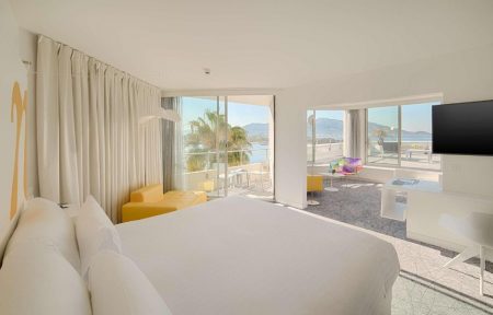 Suite d'hôtel pour séminaire en bord de mer, avec un grand lit blanc au premier plan à gauche, une méridienne jaune à côté d'un balcon vitré au centre, un bureau blanc avec une télévision murale à droite, et un espace salon donnant sur la mer et les montagnes en arrière-plan.