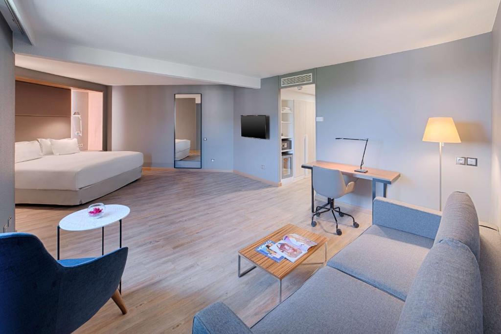 General view of a hotel room for a seminar with light wood flooring, featuring a grey sofa and low coffee table in the foreground on the right, a blue armchair and round side table on the left, a desk with chair and floor lamp on the right, a wall-mounted television in the centre, and a double bed in an alcove at the back with grey walls.