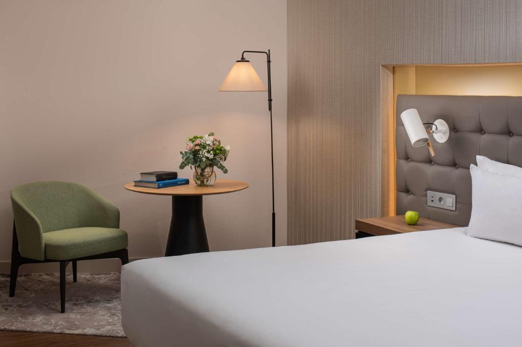 Chambre d'hôtel avec lit aux draps blancs et tête de lit rembourrée grise équipée d'une liseuse murale à droite, fauteuil rembourré vert près d'une table d'appoint ronde en bois avec bouquet de fleurs et livres, et lampadaire entre les deux à gauche, une chambre adaptée à un hôtel pour séminaire.