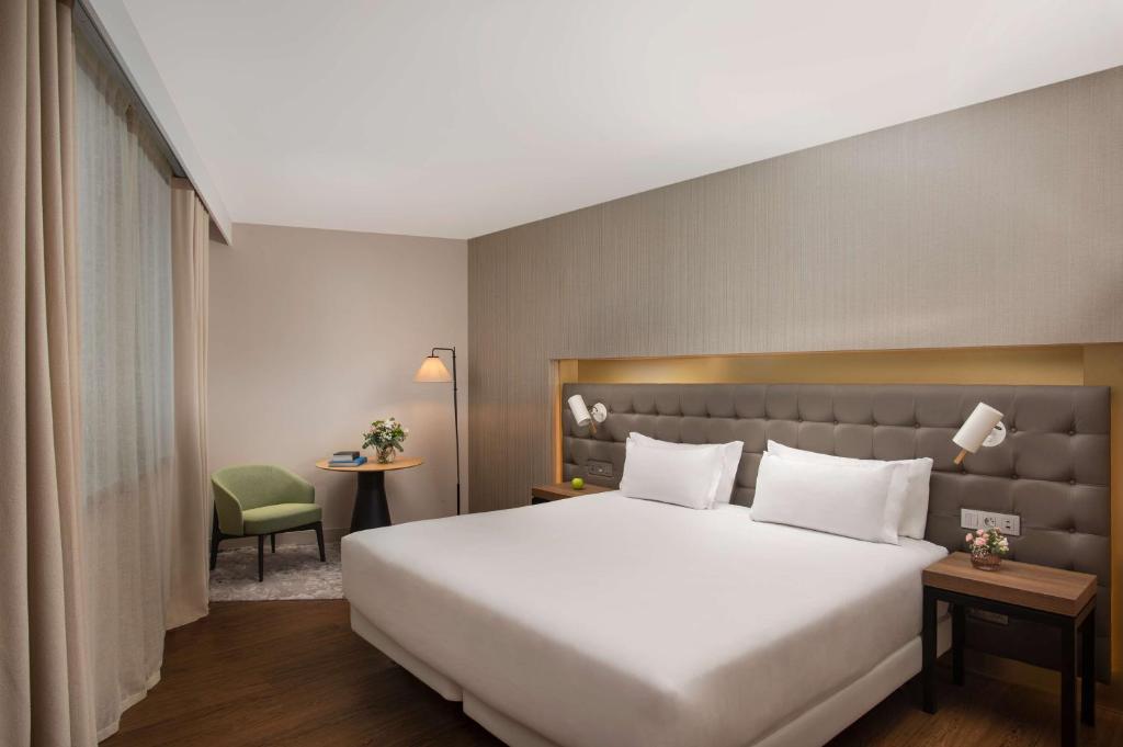 Chambre d'hôtel avec lit double aux draps blancs et tête de lit rembourrée grise équipée de deux liseuses murales au premier plan, tables de chevet en bois avec fleurs de part et d'autre, et fauteuil rembourré vert près d'une table d'appoint ronde et d'un lampadaire près de fenêtres rideautées à gauche, une chambre adaptée à un hôtel pour séminaire.