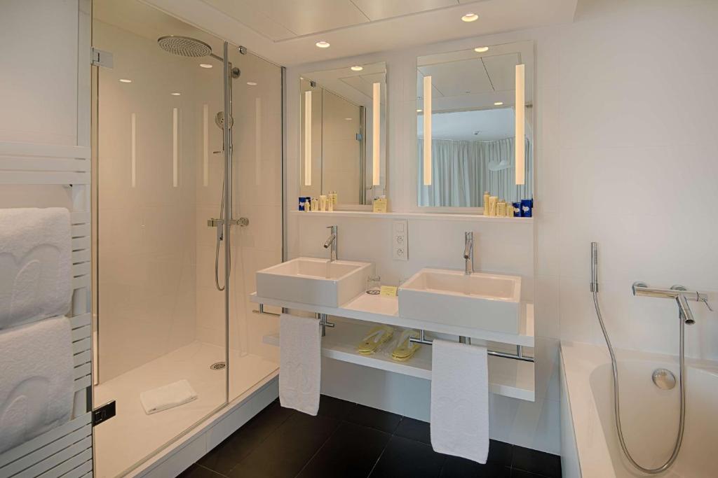 Salle de bain d'hôtel pour hébergement groupe, avec une douche à l'italienne vitrée à gauche, une double vasque avec lavabos carrés et miroirs lumineux au centre, une baignoire blanche à droite, et un sol carrelé foncé contrastant avec des murs blancs.
