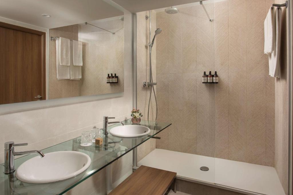 Salle de bain d'hôtel avec double vasque en verre portant deux vasques rondes et grand miroir au-dessus au premier plan, serviettes pliées reflétées dans le miroir, douche à l'italienne vitrée avec flacons de courtoisie et banc en bois à droite, adaptée à un hôtel pour séminaire.
