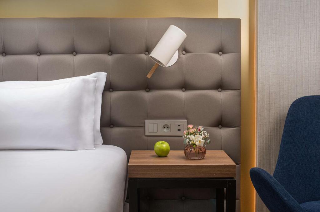 Gros plan sur un lit d'hôtel avec oreiller blanc contre une tête de lit rembourrée grise équipée d'une liseuse murale, table de chevet en bois portant une pomme verte et un petit vase de fleurs au premier plan, et fauteuil rembourré bleu visible à droite, un détail adapté à un hôtel pour séminaire.