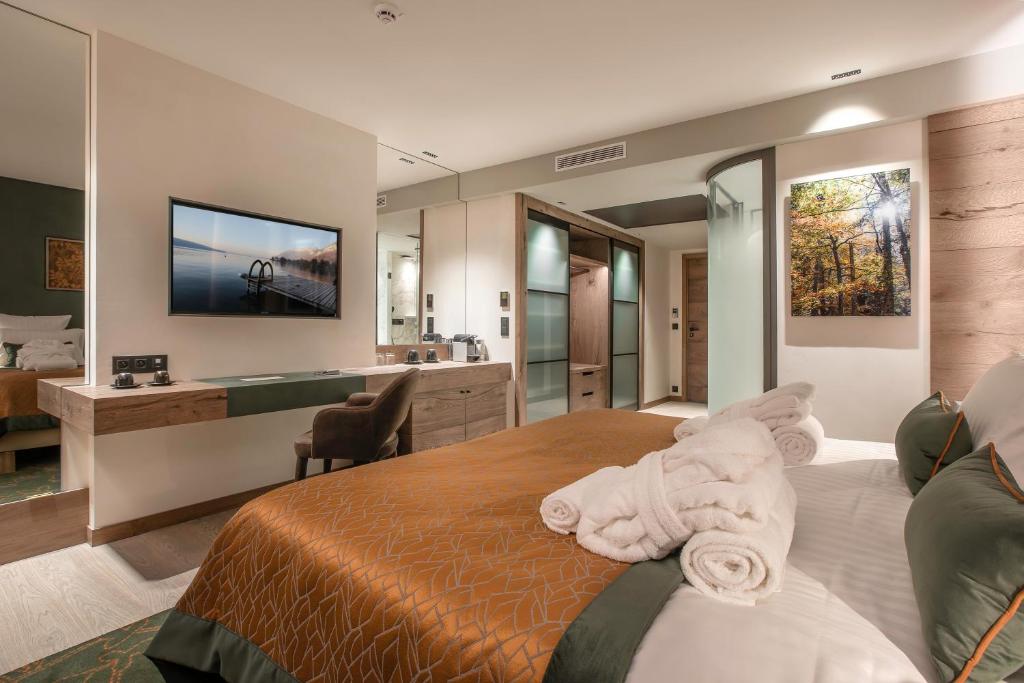 Hotel group accommodation bedroom with a large bed and white linens in the foreground, rolled towels on a brown bed runner, a wooden desk with chair on the left, and mirrored walls, wardrobe and framed forest artwork in the background.