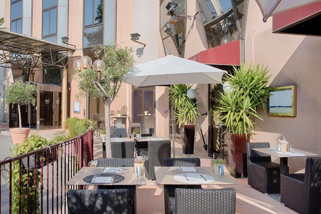 Hotel street-side terrace with dark rattan chairs and light-toned tables set with placemats and cutlery in the foreground, a large white parasol and tall potted plants in the middle ground, an olive tree and dark red metal railing on the left, and the building's glass entrance and awning in the background, tables arranged for on-site dining in a city seminar setting.