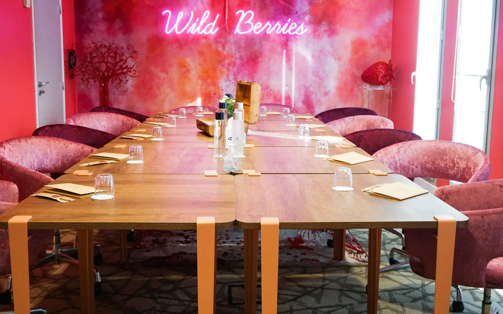 Meeting room with a long wooden table on orange legs flanked by pink velvet armchairs, glasses, notepads and toiletry items arranged along the table in the foreground, a red and orange abstract mural with a tree motif and a large strawberry sculpture behind, and a neon sign reading "Wild Berries" mounted on the wall above.