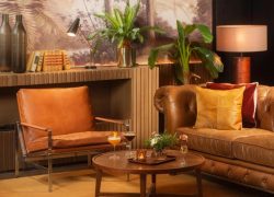 Salon d'hôtel avec fauteuil en cuir fauve et canapé chesterfield aux coussins orange et bordeaux autour d'une table basse ronde en bois dressée de cocktails au premier plan, vases décoratifs, livres et lampe de bureau sur un buffet en bois en arrière, plantes en pot hautes et fresque murale de jungle au milieu, et lampe de table à droite, un salon adapté à un afterwork.
