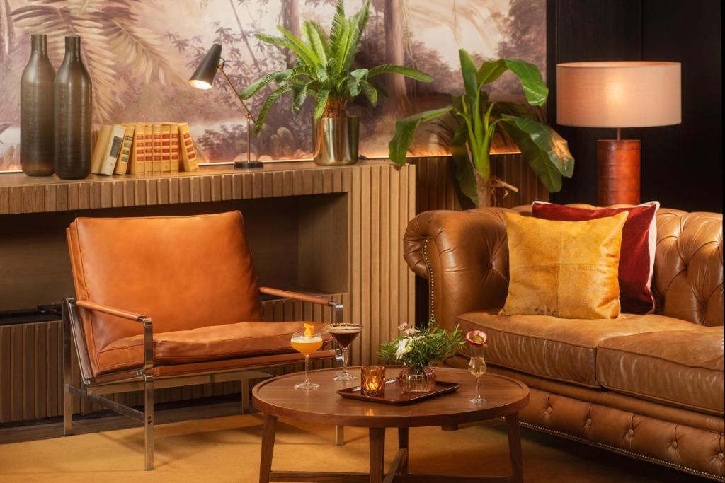 Salon d'hôtel avec fauteuil en cuir fauve et canapé chesterfield aux coussins orange et bordeaux autour d'une table basse ronde en bois dressée de cocktails au premier plan, vases décoratifs, livres et lampe de bureau sur un buffet en bois en arrière, plantes en pot hautes et fresque murale de jungle au milieu, et lampe de table à droite, un salon adapté à un afterwork.