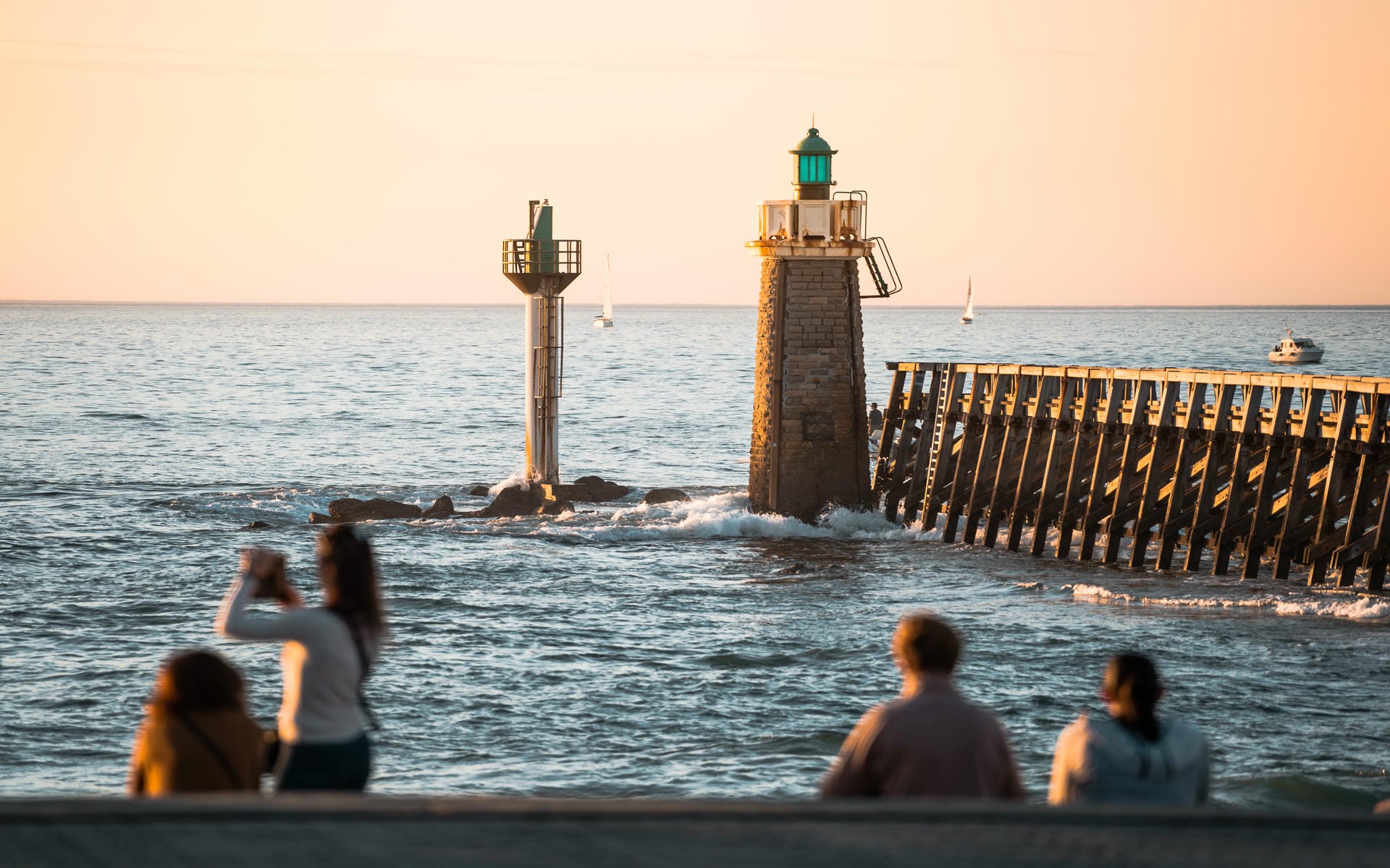Vue de l'entrée du port de Capbreton au coucher du soleil avec deux personnes photographiant la mer au premier plan, silhouettes de spectateurs assis sur un muret au premier plan en contrebas, phare en pierre et petit balise blanche signalant l'estacade en bois au milieu, voiliers et bateau à moteur visibles sur l'eau, et ciel aux tons chauds au-dessus, une scène côtière adaptée à un séminaire en bord de mer.