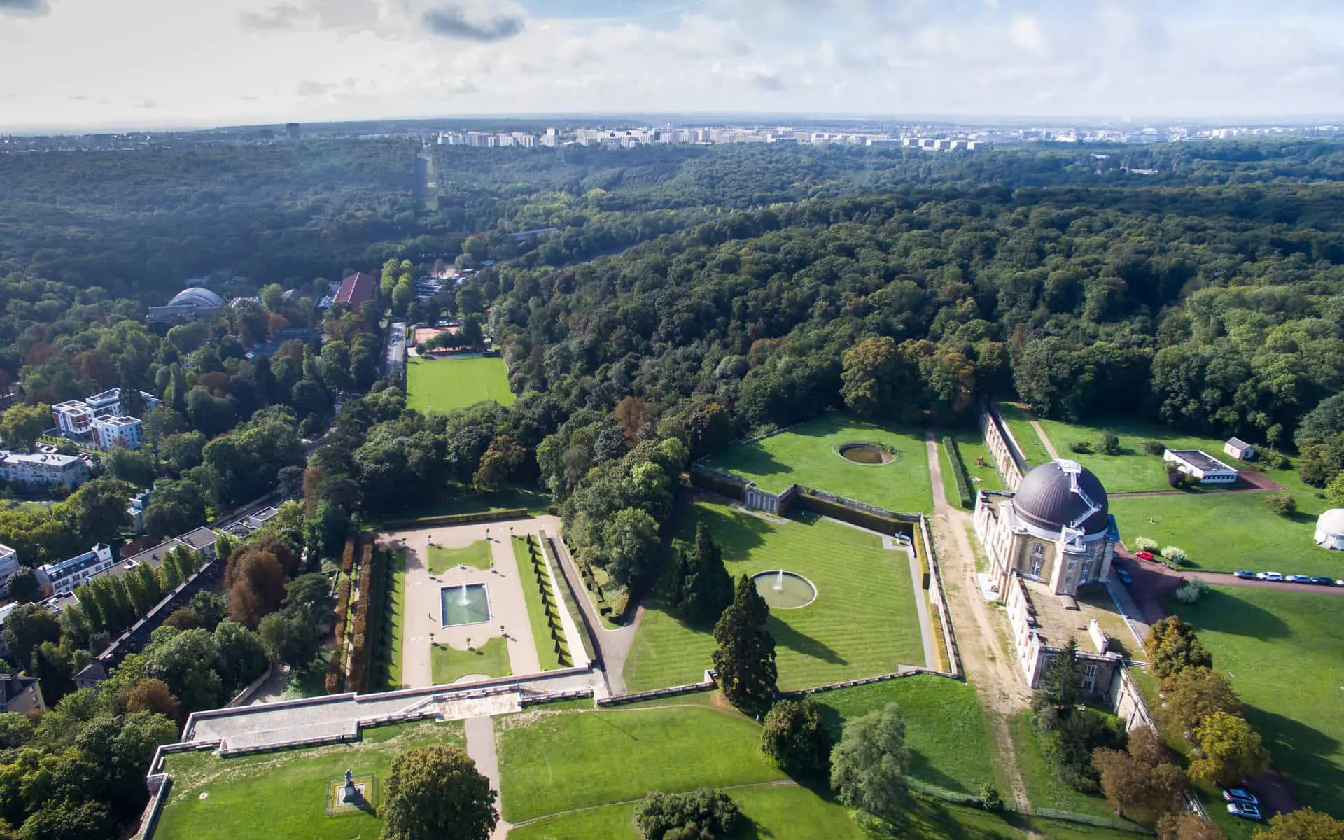 Vue aérienne d'ensemble du domaine du château de Meudon, pour un séminaire au vert, montrant un bâtiment d'observatoire à coupole avec jardins à la française, pelouses symétriques et bassins circulaires au premier plan, une forêt dense entourant le domaine, et un paysage urbain lointain à l'horizon sous un ciel partiellement nuageux.