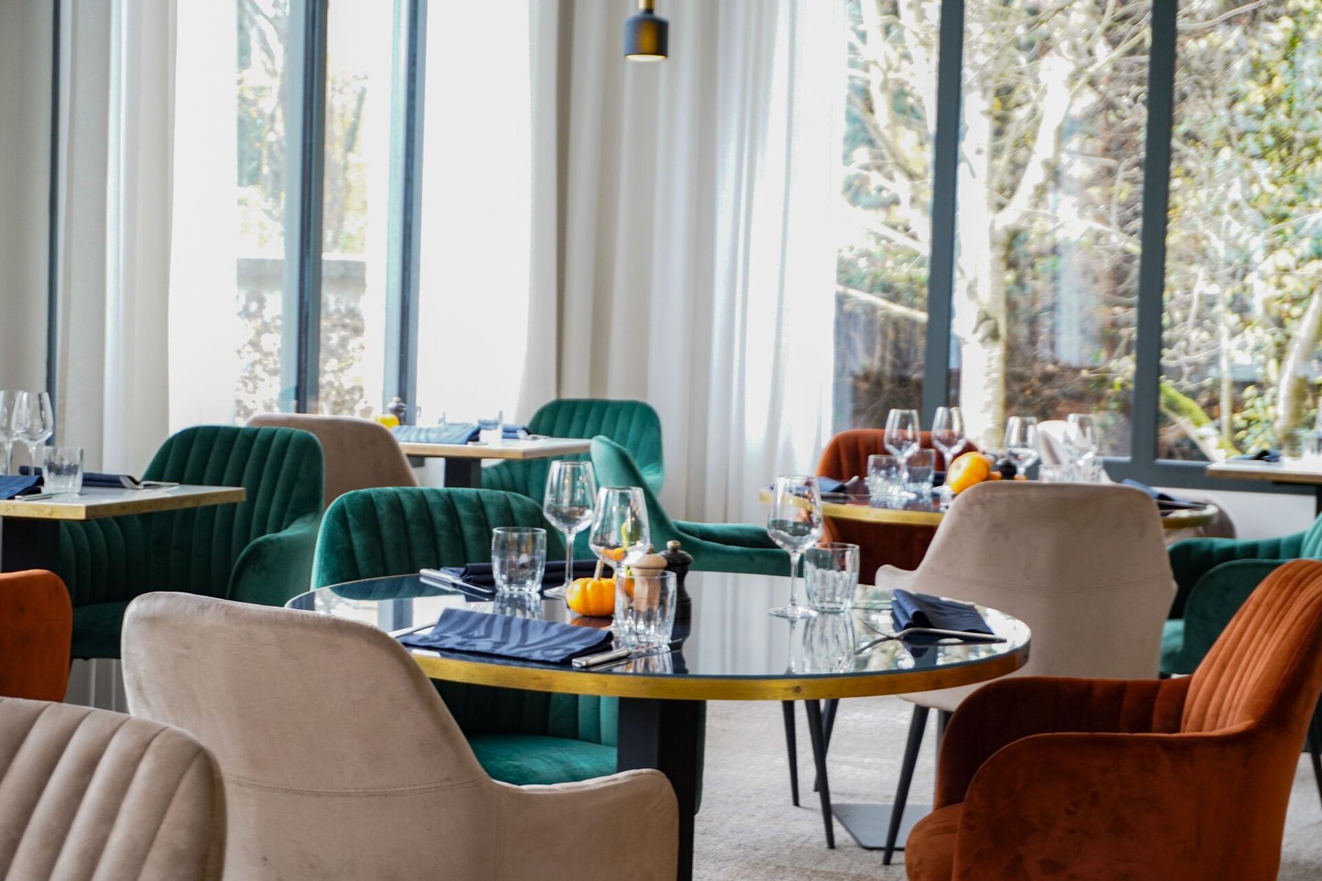 Restaurant dining room for on-site catering with a round glass table edged in gold in the foreground, set with wine glasses, navy napkins and small orange pumpkins, surrounded by teal, beige and terracotta velvet armchairs, and tall curtained windows revealing bare trees outside in the background.