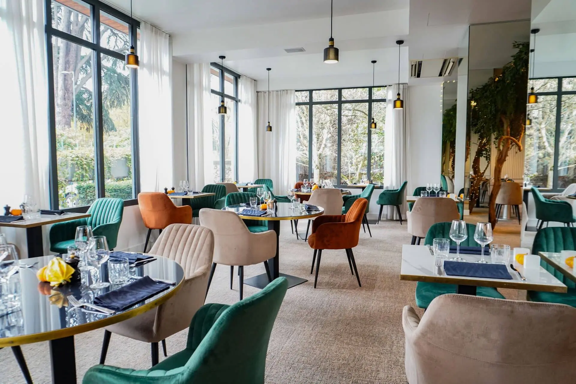 Wide view of a restaurant dining room for on-site catering with a beige carpeted floor, several round marble-topped tables set with wine glasses and navy napkins in the foreground, teal, terracotta and beige velvet chairs arranged throughout, black pendant lights hanging from a white ceiling, and floor-to-ceiling windows with sheer curtains revealing trees outside in the background.