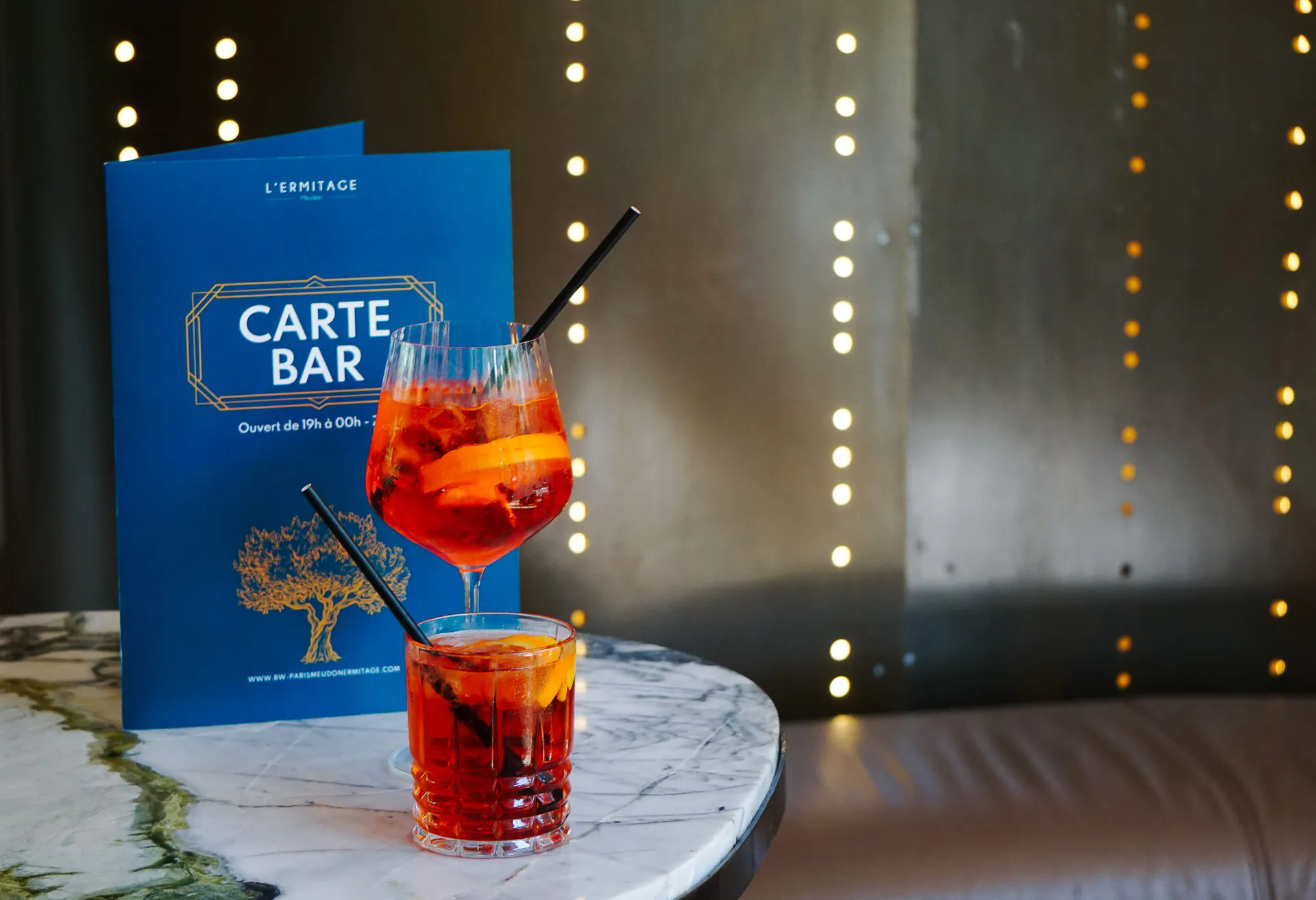 Close-up shot for an after-work event with two orange cocktails garnished with citrus slices and black straws on a white marble table in the foreground, a blue "Carte Bar" menu standing behind them, and a dark wall with vertical strings of warm lights blurred in the background.