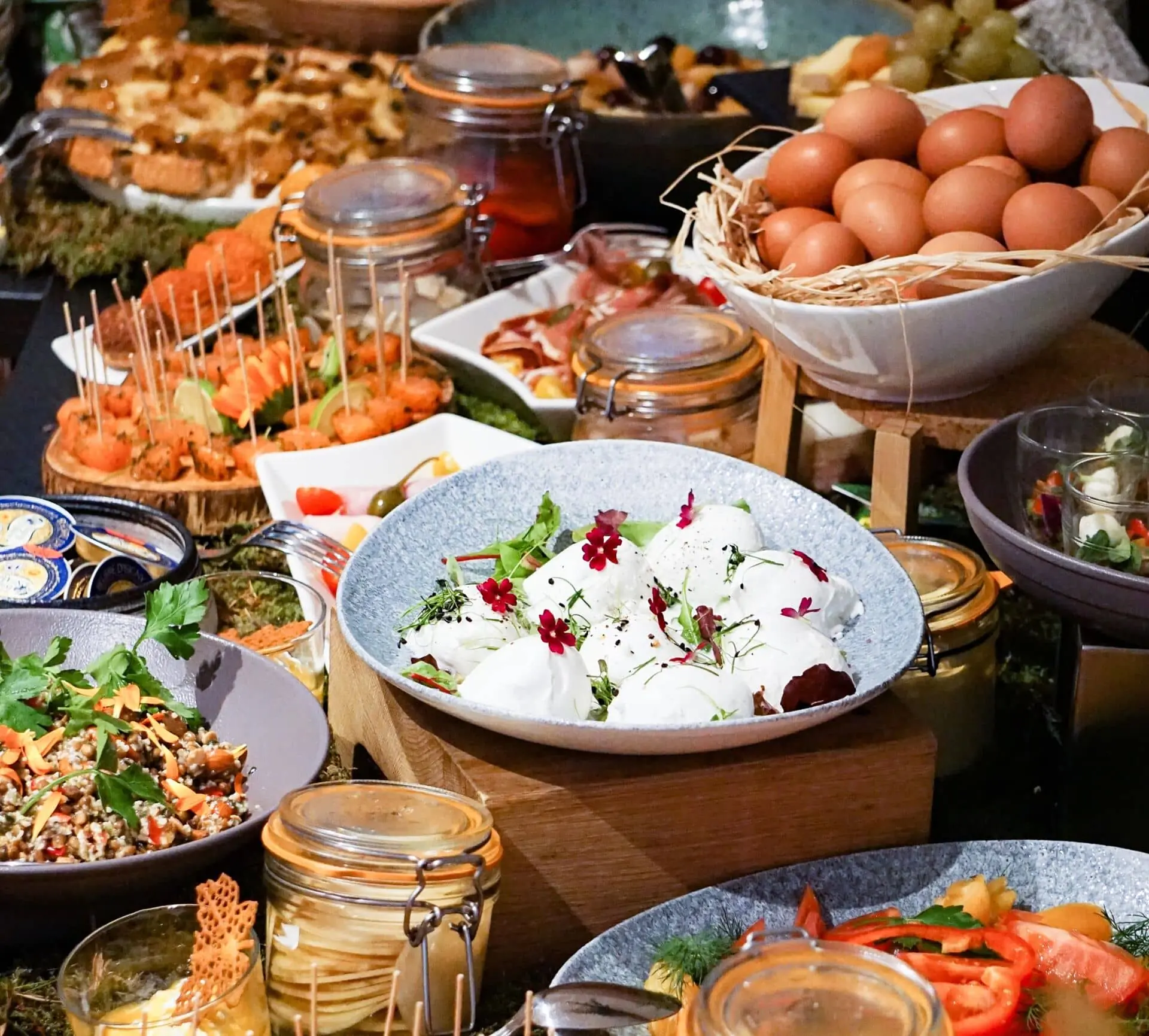 Close-up of an on-site catering buffet spread, featuring a grey bowl of burrata garnished with edible flowers and herbs on a wooden stand in the foreground, a white bowl of brown eggs nestled in straw to the right, glass jars of preserves, skewered appetisers on wooden boards, a bowl of grain salad with herbs to the left, and various dishes of charcuterie, vegetables and salads surrounding the scene.