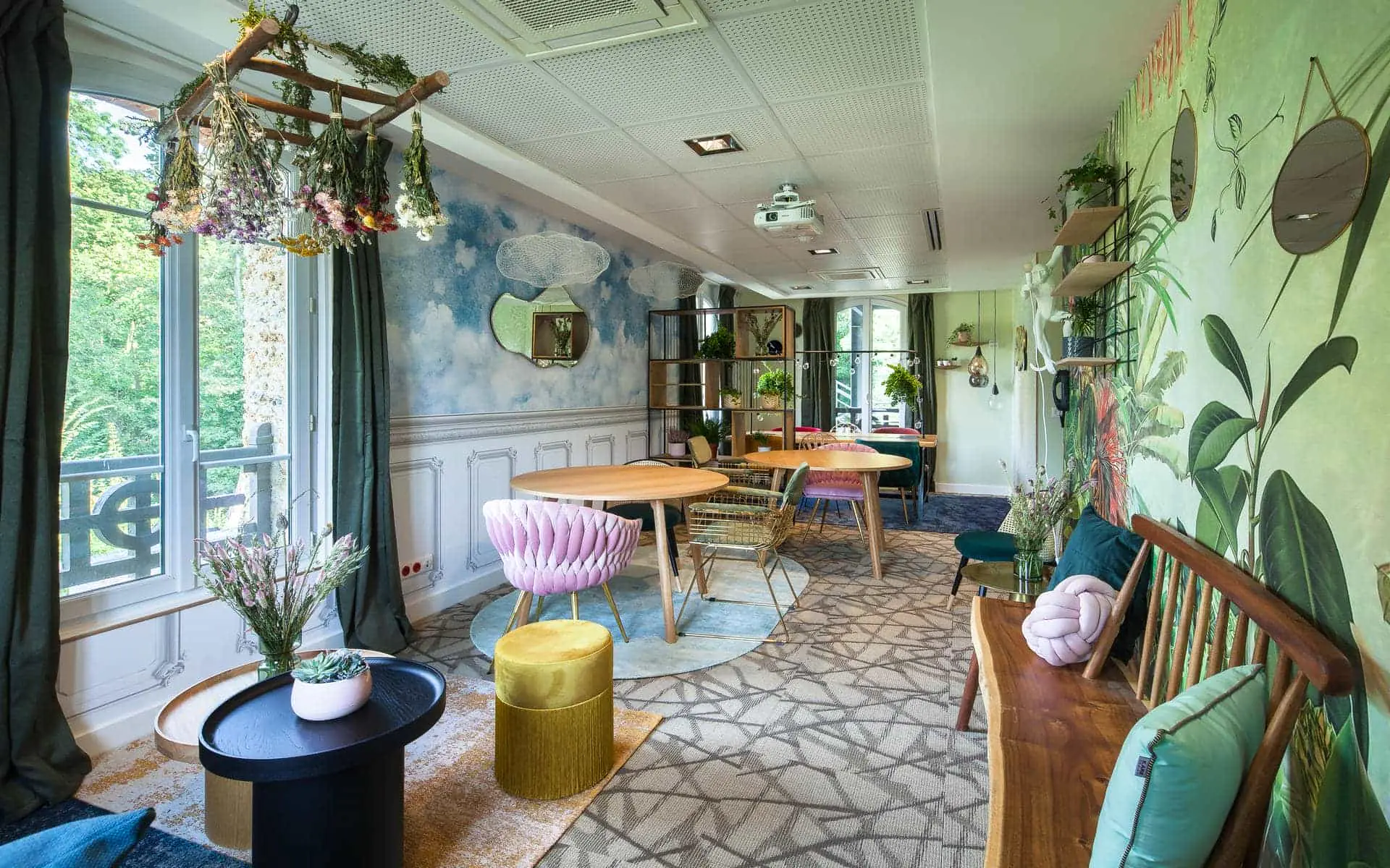 Meeting room for a séminaire bien-être with round wooden tables and pink, gold and green mismatched chairs on a patterned carpet in the foreground, a wooden bench with green cushions and a botanical-print wall on the right, an open shelving unit with plants and a cloud-shaped mirror against a sky-painted wall in the background, and a dried flower arrangement hanging from the ceiling on the left.