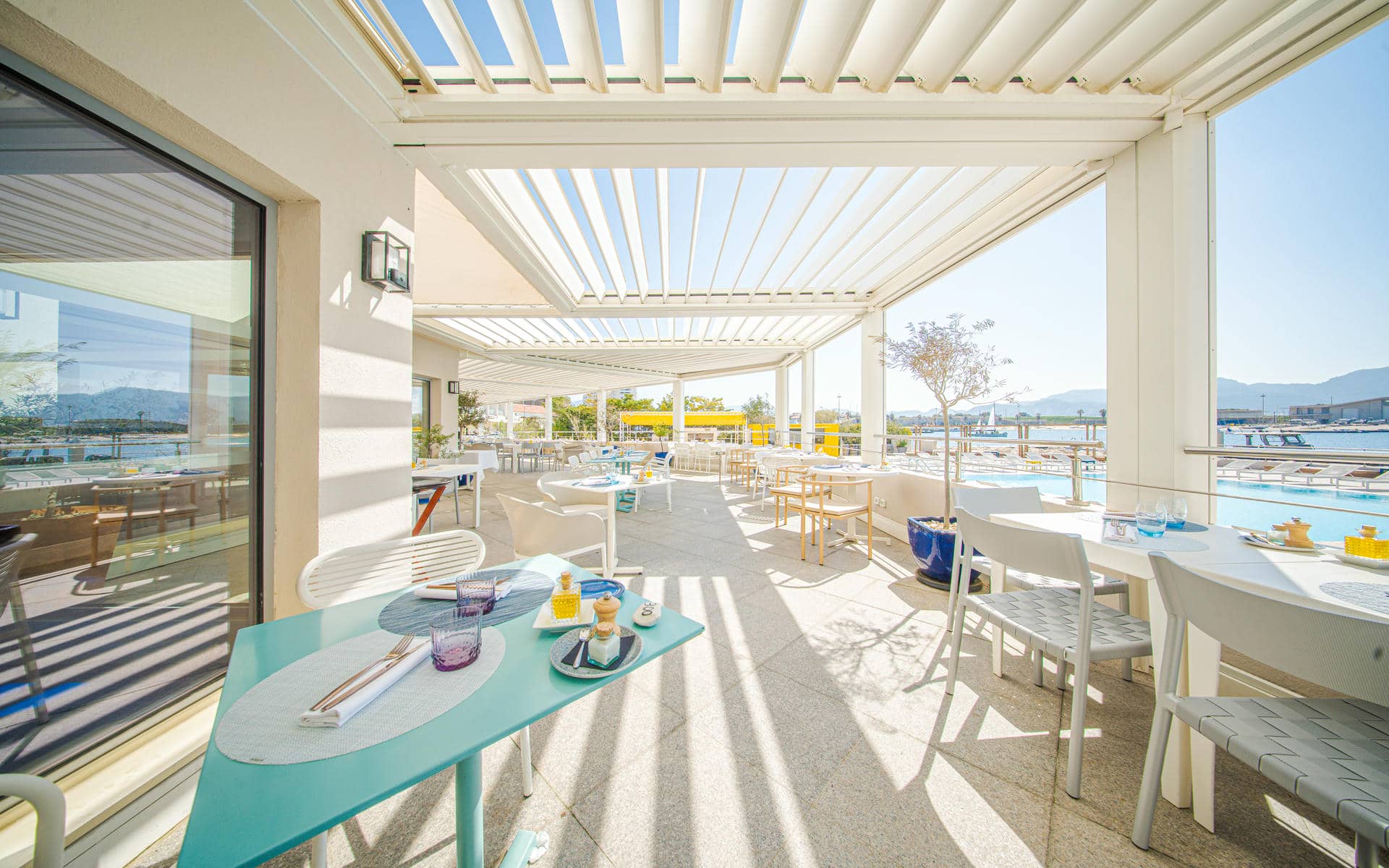 Terrasse de restaurant d'hôtel pour restauration sur place en vue générale avec des tables turquoise et blanches dressées de vaisselle, verres et condiments au premier plan, des chaises blanches réparties sur toute la terrasse, une pergola blanche au-dessus créant des ombres rayées sur le sol, et un port de plaisance avec des montagnes à l'arrière-plan à droite.