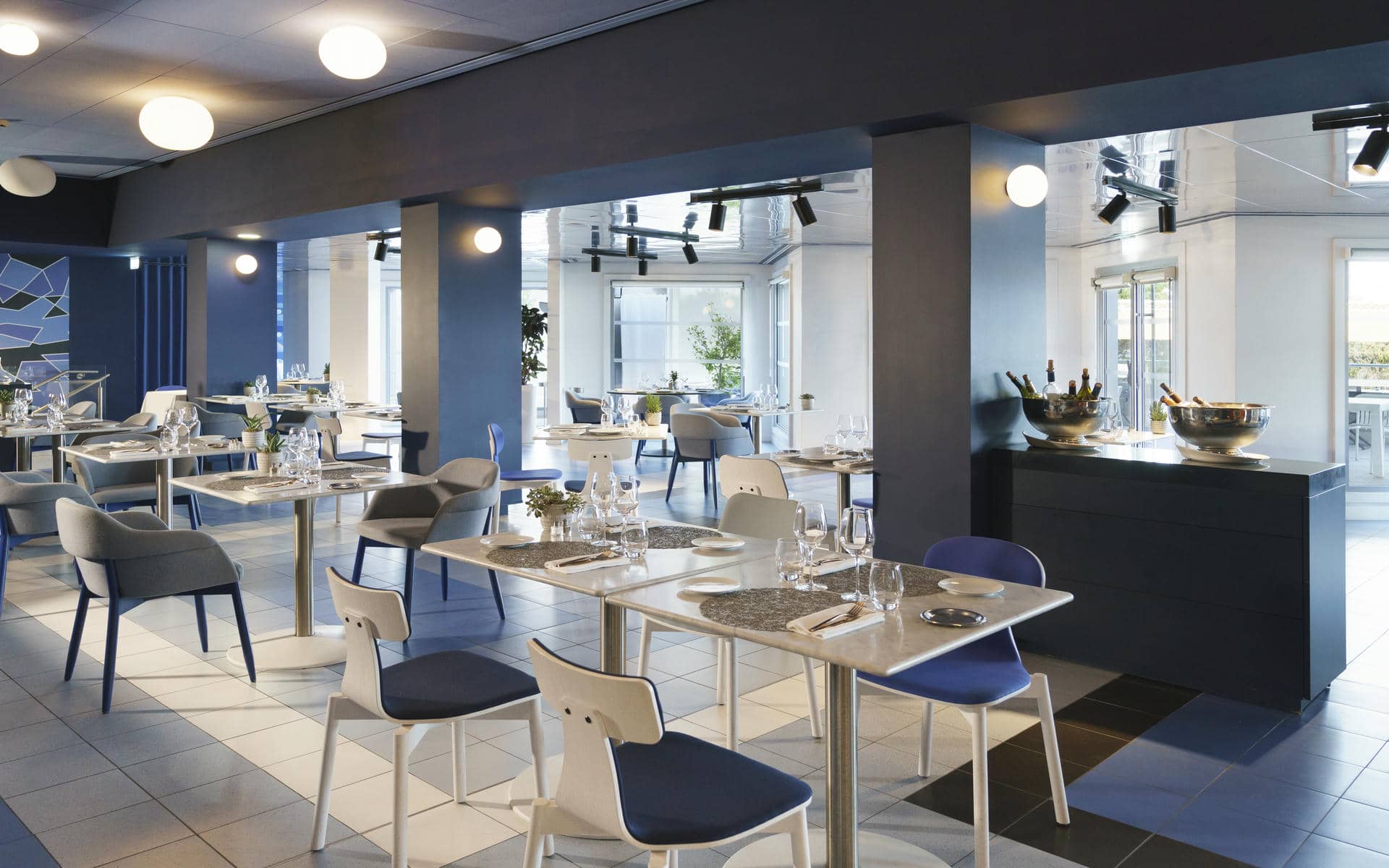 Restaurant d'hôtel pour restauration sur place en vue générale avec des tables carrées dressées de vaisselle blanche, verres et couverts au premier plan, des chaises bleues et grises réparties dans la salle, un buffet noir avec des seaux à glace à droite, de grandes fenêtres à l'arrière-plan, et un sol en carrelage gris.