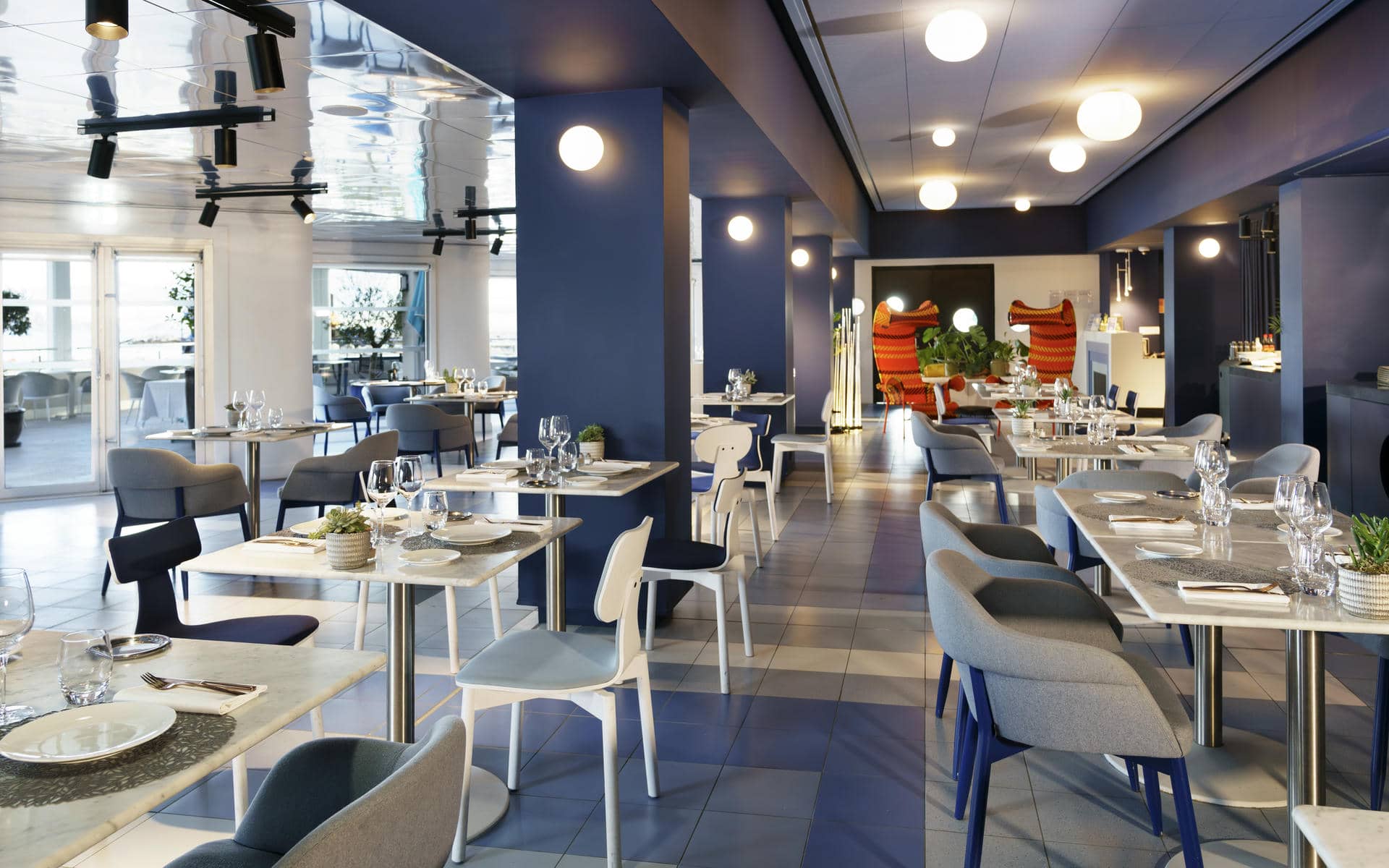 Restaurant d'hôtel pour restauration sur place en vue générale avec des tables carrées dressées de vaisselle blanche, verres et couverts au premier plan, des chaises bleues et grises réparties dans la salle, des colonnes bleues au centre, de grandes baies vitrées donnant sur une terrasse à gauche, et un sol en carrelage gris.