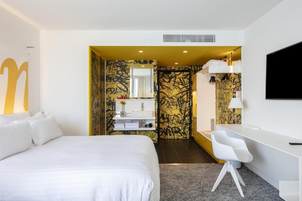 Chambre d'hôtel pour hébergement groupe, avec un grand lit blanc à gauche au premier plan, un bureau blanc avec chaise et une télévision murale à droite, et un espace salle de bain ouvert avec une vasque blanche, un plafond jaune et un mur à motifs graphiques noirs en arrière-plan sur un sol carrelé foncé.