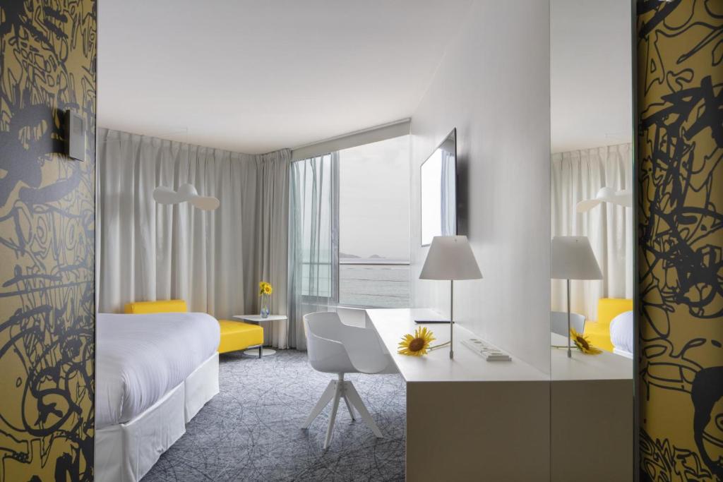 Chambre d'hôtel pour séminaire en bord de mer, avec un grand lit blanc à gauche au premier plan, un bureau blanc avec chaise et une télévision murale à droite, une méridienne jaune à côté d'une grande fenêtre d'angle au centre, et une vue sur la mer en arrière-plan sur une moquette grise à motifs.