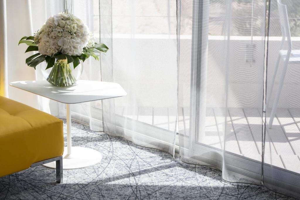 Chambre d'hôtel pour séminaire au vert avec un canapé jaune à gauche, une table d'appoint blanche accueillant un vase en verre avec des fleurs blanches au centre, des rideaux voilés toute hauteur devant une porte vitrée à droite, et une moquette à motif graphique au premier plan.