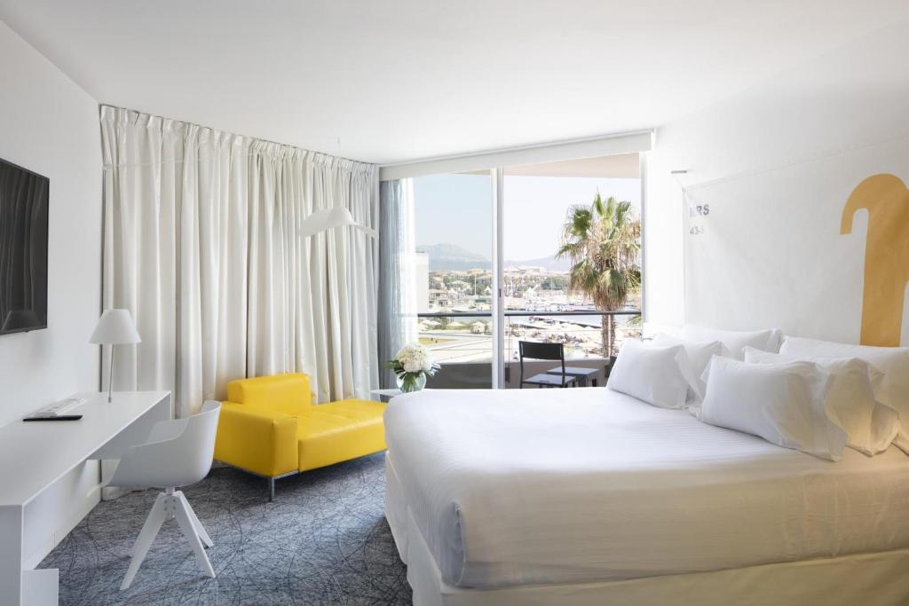 Chambre d'hôtel pour séminaire en bord de mer, avec un grand lit blanc à droite au premier plan, un bureau blanc avec chaise à gauche, un fauteuil jaune à côté d'un lampadaire au centre, et un balcon donnant sur des palmiers, le port de plaisance et les montagnes en arrière-plan.