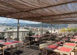 Terrasse sur le toit d'un hôtel ombragée par une pergola en roseau avec guirlande d'ampoules suspendues, tables grises aux chaises à accoudoirs en bois dressées de sets de table rouges et petits bouquets de fleurs au premier plan, arbres en pot et murets bordant la terrasse à droite, et collines et toits de la ville de Nice visibles en arrière-plan, tables disposées pour une restauration sur place dans un cadre de séminaire en ville.