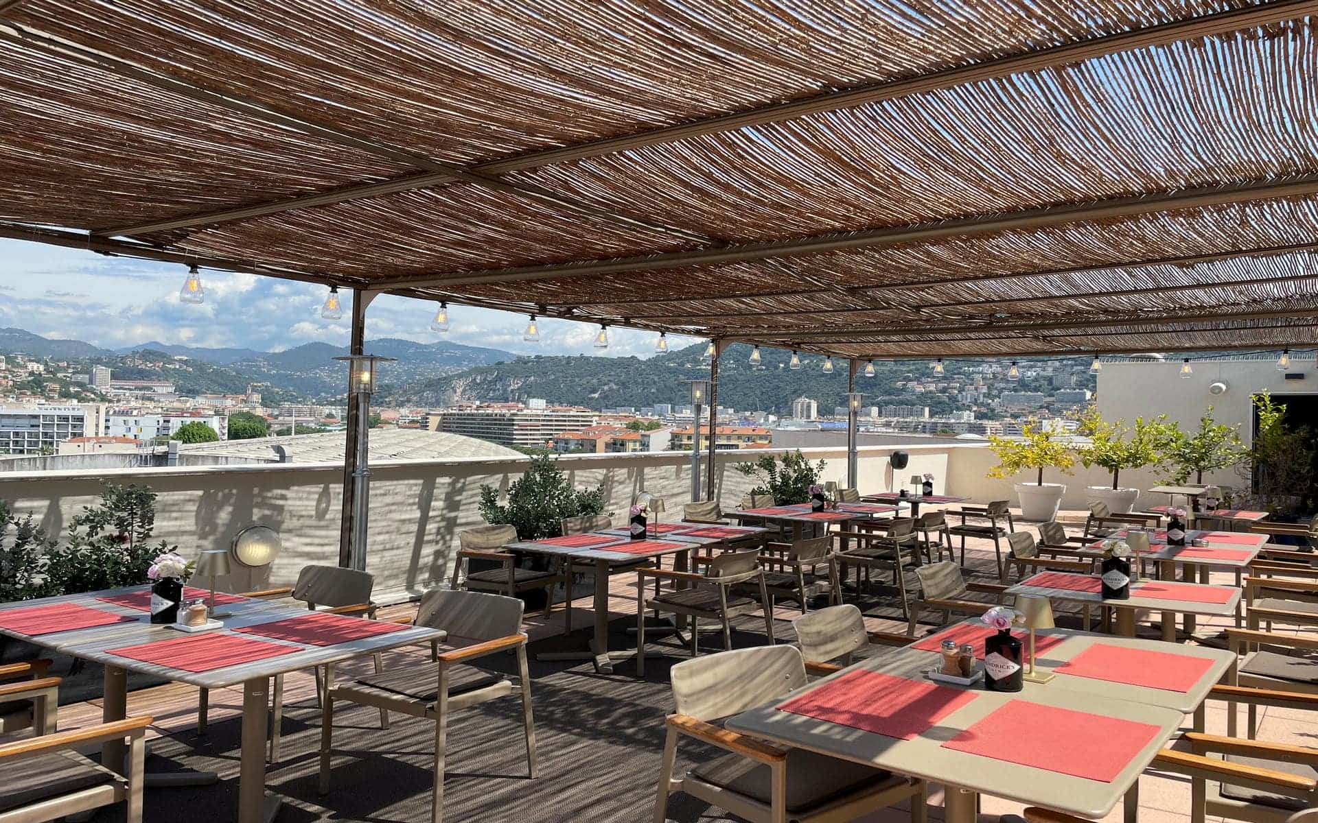 Hotel rooftop terrace shaded by a reed pergola with hanging bulb lights, grey tables with wooden-armed chairs set with red placemats and small flower arrangements in the foreground, potted trees and low walls bordering the terrace on the right, and the hills and rooftops of Nice visible in the background, tables arranged for on-site dining in a city seminar setting.