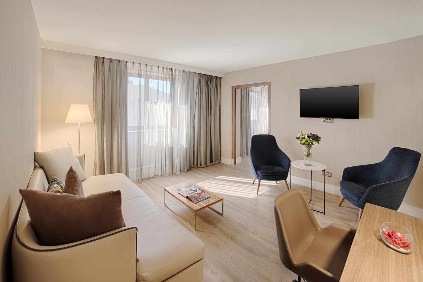 Hotel suite living area for group accommodation with a beige sofa and floor lamp on the left, a light wood coffee table in the centre, two dark blue armchairs and a round side table with flowers beneath a wall-mounted television on the right, large windows with sheer curtains at the back, and light wood flooring.