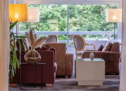Espace lounge avec des fauteuils et canapés en cuir marron, de grands lampadaires allumés et des rideaux encadrant l’entrée du bar, qui se prolonge vers la salle de restaurant dotée de grandes baies vitrées donnant sur la nature.