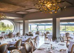 Restaurant de séminaire avec de grandes baies vitrées et une large fenêtre ronde donnant sur le domaine, des plafonniers en forme de feuilles dorées, des chaises beiges en bois et tissu, et des tables carrées.
