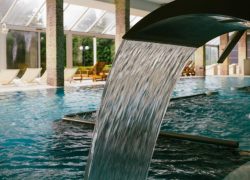 Bec d’eau dans la piscine intérieure de l’hôtel, avec un espace balnéo ou bain bouillonnant et des transats disposés devant de grandes baies vitrées donnant sur la nature.