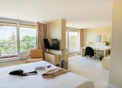 Suite avec un balcon aménagé donnant sur le domaine, comprenant un espace chambre et un espace salon dans des tons clairs, avec climatisation et mobilier blanc.